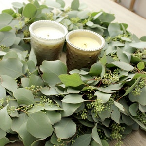 May include: Two candles in glass holders with a textured surface sit on a bed of eucalyptus greenery. The candles are white and gold.