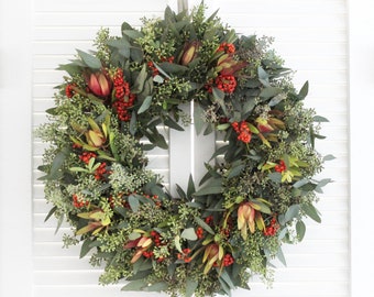 May include: A green, red, and brown wreath made of eucalyptus, berries, and flowers. The wreath is hanging on a white door.