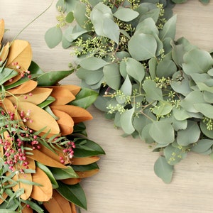 May include: Two wreaths made of natural greenery. One wreath is made of magnolia leaves and has red berries. The other wreath is made of eucalyptus leaves.