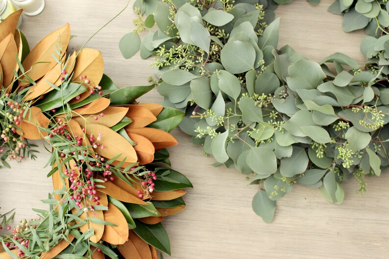 May include: Two wreaths made of natural greenery. One wreath is made of magnolia leaves and has red berries. The other wreath is made of eucalyptus leaves.