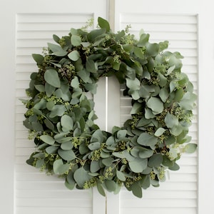 May include: A green eucalyptus wreath with small green buds. The wreath is hanging on a white wooden door.