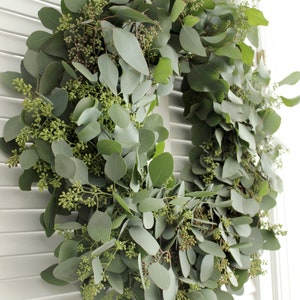 May include: A green eucalyptus wreath with small green buds, hanging on a white wooden door.
