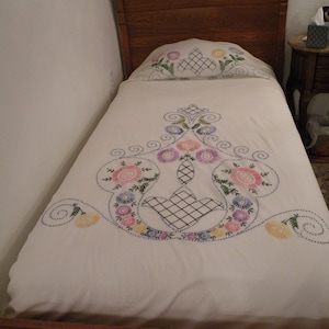 May include: A white bedspread with embroidered floral designs in shades of pink, purple, green, and yellow. The bedspread is on a wooden bed frame with a headboard.