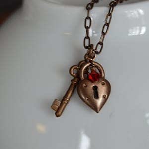 May include: A bronze heart-shaped padlock necklace with a key charm and a red crystal accent. The necklace is on a white surface.