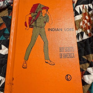 May include: An orange book titled "Indian Lore" by Boy Scouts of America. The cover features an illustration of a person in a scout uniform with a backpack. The person is wearing a red hat and carrying a large red and brown backpack.