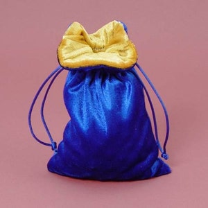 May include: A royal blue velvet drawstring bag with a gold-colored interior and drawstring. The bag is gathered at the top with a drawstring closure. The background is a solid pink color.