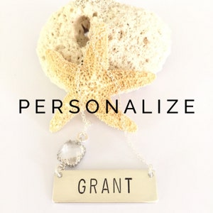 May include: A silver necklace with a clear crystal charm and a personalized nameplate that says "GRANT". The necklace is displayed on a white background with a starfish.