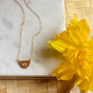 May include: A gold chain necklace with a small, half-circle pendant featuring a sun design. The necklace is displayed on a white marble surface with a yellow flower in the background.