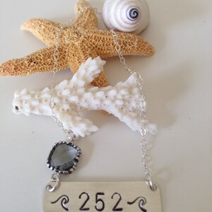 May include: Silver necklace with a silver bar pendant that says "252" with a wave design on either side. The pendant is attached to a silver chain with a dark gray gemstone charm.