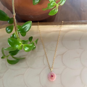 May include: A delicate gold necklace featuring a pink flower pendant with a small red center stone. The necklace hangs on a gold chain. The image is set against a neutral background with a green plant in the upper left corner.