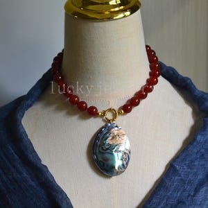 May include: A beaded necklace with a large oval pendant. The necklace features red beads and a gold clasp. The pendant is an iridescent abalone shell with gold accents. The text "lucky jet" is visible on the necklace.
