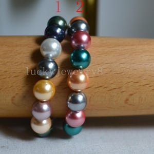 Op de afbeelding: Twee armbanden met glanzende, ronde parels in verschillende kleuren. De eerste armband heeft donkergroene, witte en gouden parels. De tweede armband heeft grijze, rode en turquoise parels. De tekst "lucky jewelry8" is zichtbaar.