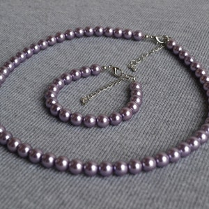 May include: A matching necklace and bracelet set featuring lavender-colored faux pearls. The necklace and bracelet are strung together with a silver-colored chain and clasp. The pearls are evenly spaced and have a smooth, glossy finish.