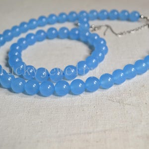 May include: A light blue beaded necklace with a silver clasp and chain. The necklace is made of round, smooth beads. The beads are a soft, pastel blue color. The necklace is displayed on a white surface.
