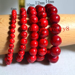 Peut inclure: Plusieurs bracelets de perles rouges de différentes tailles : 6mm, 8mm, 10mm, 12mm et 14mm. Les perles présentent un aspect marbré. Les bracelets sont présentés sur une cheville en bois.