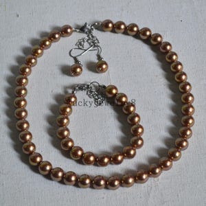 May include: A set of brown pearl jewellery featuring a necklace, bracelet, and earrings. The necklace and bracelet are made of round pearls strung on a silver chain. The earrings are simple studs with a single pearl.