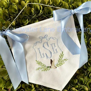 May include: A white fabric banner with a blue ribbon tied at the top. The banner features a monogram of the letters "LSE" in a decorative frame with a bird and green leaves. The text "Sweetwater Lane Embroidery" is visible at the top of the banner.