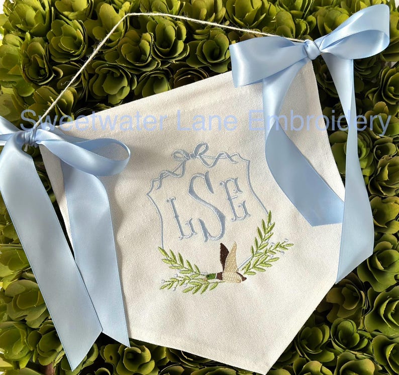 May include: A white fabric banner with a blue ribbon tied at the top. The banner features a monogram of the letters "LSE" in a decorative frame with a bird and green leaves. The text "Sweetwater Lane Embroidery" is visible at the top of the banner.