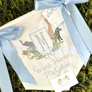 Peut inclure: Une banni&egrave;re en toile blanche avec des n&oelig;uds de ruban bleu, pr&eacute;sentant un monogramme brod&eacute; "M" entour&eacute; d'une couronne de feuilles vertes et d'illustrations d'animaux. La banni&egrave;re comprend &eacute;galement le texte brod&eacute; "Grant Harvey McGhee".