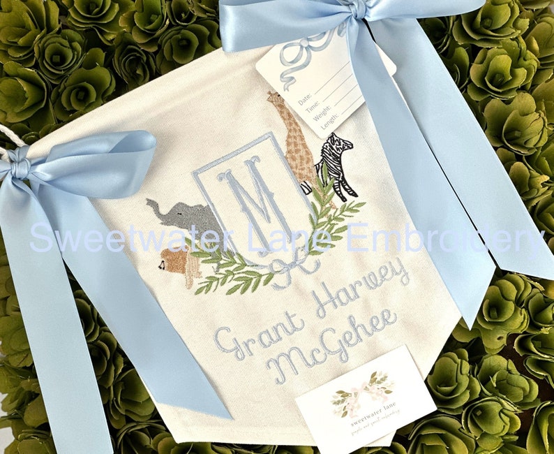 Peut inclure: Une banni&egrave;re en toile blanche avec des n&oelig;uds de ruban bleu, pr&eacute;sentant un monogramme brod&eacute; "M" entour&eacute; d'une couronne de feuilles vertes et d'illustrations d'animaux. La banni&egrave;re comprend &eacute;galement le texte brod&eacute; "Grant Harvey McGhee".