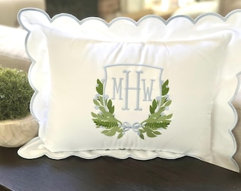 May include: A white decorative pillow with a scalloped edge and a light blue trim. The pillow features an embroidered monogram within a laurel wreath design in shades of green and light blue.