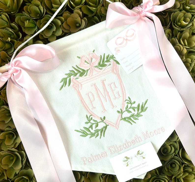 May include: A white banner with pink ribbon ties, featuring a green and pink embroidered monogram "PME" inside a shield with leafy accents. The banner also includes the text "Palmer Elizabeth Moore".