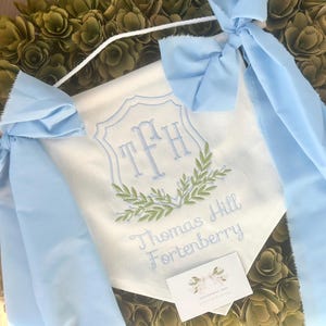 May include: A white fabric banner featuring light blue embroidered monogram letters TFH inside a crest, accented by green leaf embroidery. The banner includes the text Thomas Hill Fortenberry in light blue script and light blue fabric bows.