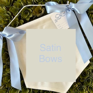 May include: A white canvas banner with the text "Satin Bows" printed in light blue. The banner is decorated with two light blue satin bows.