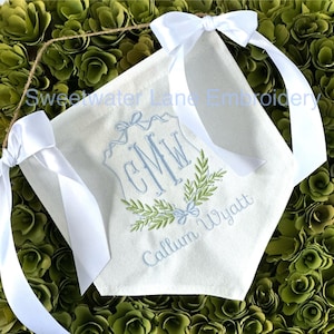 May include: A white canvas banner with a blue and green embroidered monogram "CMW" and the name "Callum Wyatt" below. The banner has white satin ribbon ties.