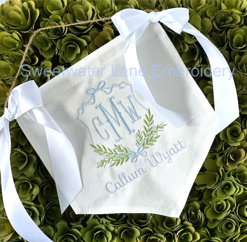 May include: A white canvas banner with a blue and green embroidered monogram "CMW" and the name "Callum Wyatt" below. The banner has white satin ribbon ties.