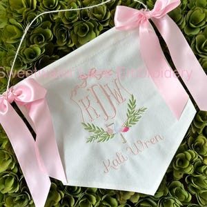 May include: A white banner with pink ribbon ties. The banner is embroidered with the monogram "KJW" and the name "Kali Wren" in pink thread. A small bird and floral design are also embroidered on the banner.