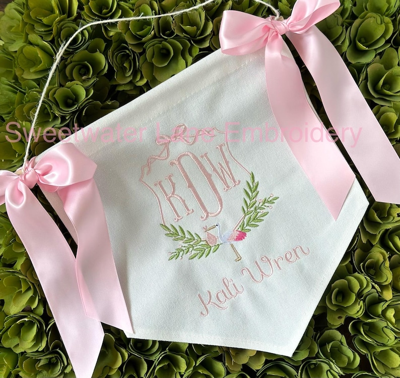 May include: A white banner with pink ribbon ties. The banner is embroidered with the monogram "KJW" and the name "Kali Wren" in pink thread. A small bird and floral design are also embroidered on the banner.