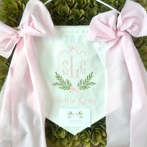 May include: A white banner with pink satin ribbons, featuring a monogram of the letters "S L G" in a shield with green leafy branches. The banner also includes the text "Scottie Gray".