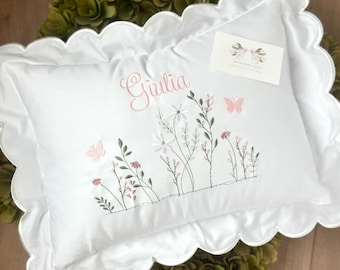 May include: White pillow with a scalloped edge, embroidered with a floral design, butterflies, and the name "Giulia" in pink script. A small card with floral graphics rests on the pillow.