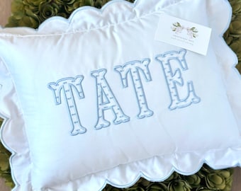 May include: White pillow with a scalloped edge and blue trim. The pillow has the name "TATE" embroidered in blue on the front.