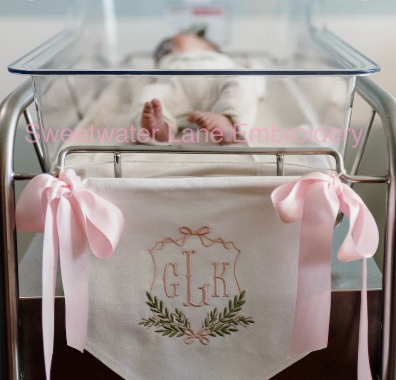 May include: A white crib banner with pink satin ribbons and a monogram embroidered in a shield shape with the initials "GLK" and a wreath of green leaves and pink bows.