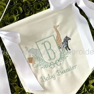 Peut inclure: Une banni&egrave;re en tissu blanc avec un monogramme brod&eacute; en vert "B" entour&eacute; d'une couronne de feuilles. La banni&egrave;re pr&eacute;sente &eacute;galement des images brod&eacute;es d'une girafe, d'un z&egrave;bre et d'un &eacute;l&eacute;phant. Le texte "Baby Bouvier" est brod&eacute; sous le monogramme.