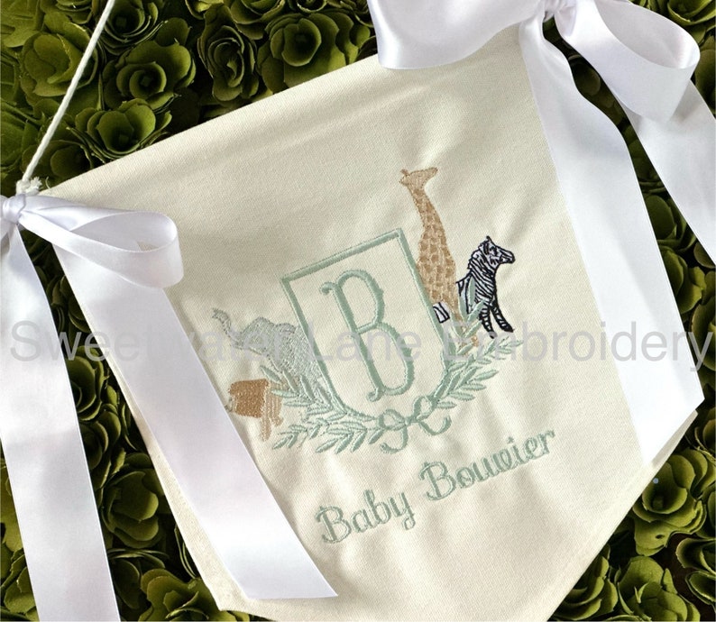 Peut inclure: Une banni&egrave;re en tissu blanc avec un monogramme brod&eacute; en vert "B" entour&eacute; d'une couronne de feuilles. La banni&egrave;re pr&eacute;sente &eacute;galement des images brod&eacute;es d'une girafe, d'un z&egrave;bre et d'un &eacute;l&eacute;phant. Le texte "Baby Bouvier" est brod&eacute; sous le monogramme.