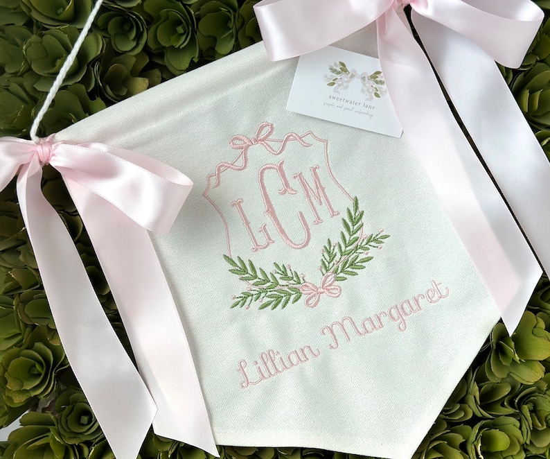 May include: A white fabric banner with pink satin ribbon ties. The banner features a monogram with the initials "LCM" in a script font, surrounded by a wreath of green leaves and pink bows. The name "Lillian Margaret" is embroidered below the monogram.