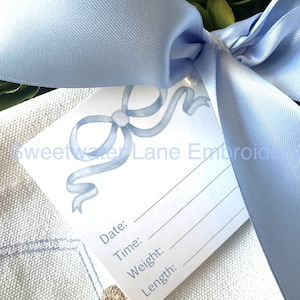 May include: A white card with a blue ribbon design and the text "Sweetwater Lane Embroidery". The card has blank lines for "Date", "Time", "Weight", and "Length". The card is tied with a light blue satin ribbon.