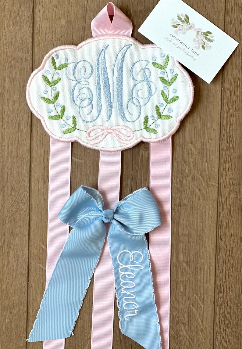 K&ouml;nnte beinhalten: Ein rosa-wei&szlig;er monogrammierter Haarbogenhalter mit drei rosa Schleifen. Ein blauer Bogen mit dem Namen "Eleanor" ist auf den Halter gestickt.