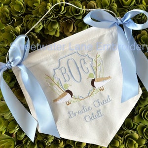 May include: A white fabric banner with blue ribbon ties. The banner features a monogram "BOC" in a blue shield with two embroidered ducks on a branch. The text "Brodie Chad Odell" is embroidered below the monogram.