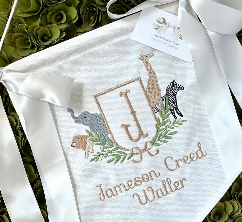 Peut inclure: Une banni&egrave;re en toile blanche avec un monogramme dor&eacute; des lettres "J" et "U" entour&eacute; d'une couronne de laurier verte. La couronne est d&eacute;cor&eacute;e d'images brod&eacute;es d'une girafe, d'un z&egrave;bre, d'un &eacute;l&eacute;phant et d'un lion. La banni&egrave;re comprend &eacute;galement le texte brod&eacute; "Jameson Creed Waller".