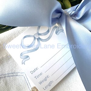 Peut inclure: Une carte blanche avec un motif de ruban bleu et le texte "Sweetwater Lane Embroidery". La carte comporte des lignes vierges pour "Date", "Heure", "Poids" et "Longueur". La carte est attach&eacute;e avec un ruban de satin bleu clair.