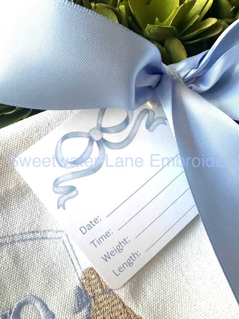 Peut inclure: Une carte blanche avec un motif de ruban bleu et le texte "Sweetwater Lane Embroidery". La carte comporte des lignes vierges pour "Date", "Heure", "Poids" et "Longueur". La carte est attach&eacute;e avec un ruban de satin bleu clair.