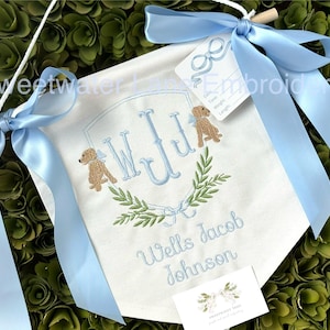 May include: A white banner with blue ribbon, embroidered with the monogram "WJJ" in blue, surrounded by green leaves and two brown dogs. The banner also reads "Wells Jacob Johnson".