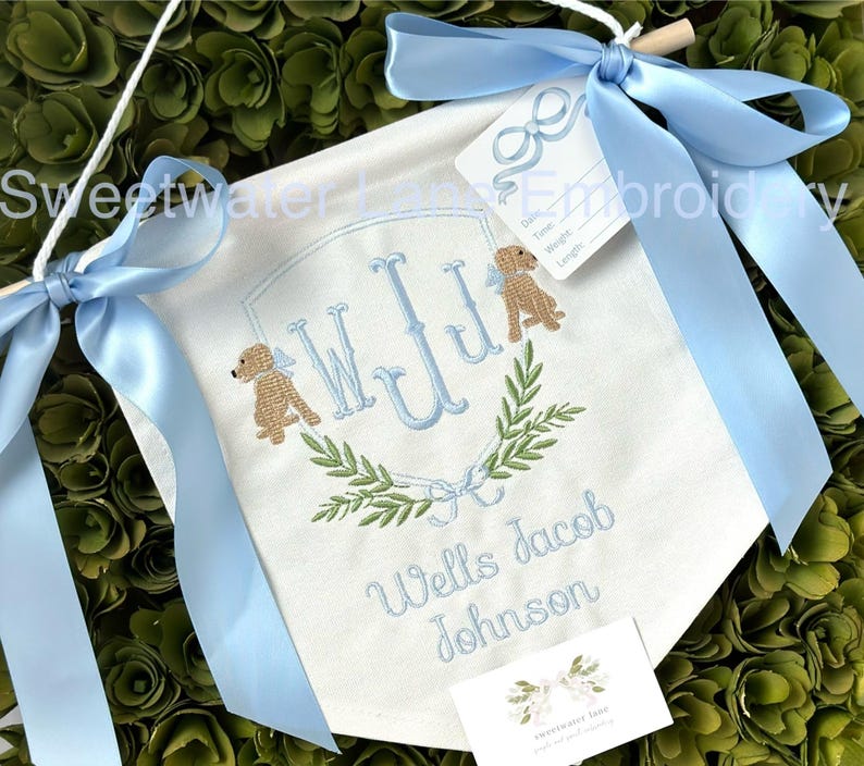 May include: A white banner with blue ribbon, embroidered with the monogram "WJJ" in blue, surrounded by green leaves and two brown dogs. The banner also reads "Wells Jacob Johnson".