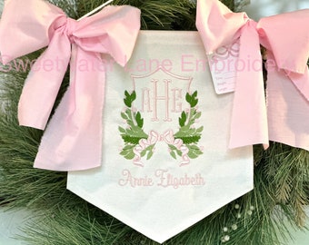 May include: A white fabric banner featuring pink ribbon bows at the top corners, an embroidered crest with the monogram MHE, green leaf and pink berry accents, and the name Annie Elizabeth in pink text. A small tag displays Date, Time, Weight, Length.