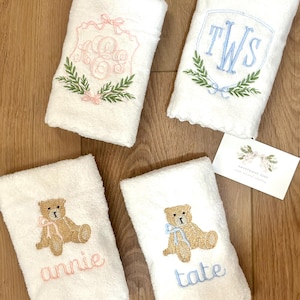 May include: Four white towels with embroidered designs. Two towels feature a teddy bear with a bow, one with a pink bow and the name "annie" embroidered below, the other with a blue bow and the name "tate" embroidered below. One towel features a monogram "TWS" with a wreath of leaves. The last towel features a monogram with a wreath of leaves and a pink bow.