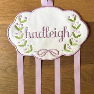 K&ouml;nnte beinhalten: Ein wei&szlig;es, wolkenf&ouml;rmiges Schild aus Stoff mit dem Namen "hadleigh", der in Lila gestickt ist. Das Schild hat eine Schleife aus lila Band zum Aufh&auml;ngen und vier lila B&auml;nder, die herunterh&auml;ngen.